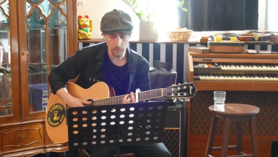 Tuomas Kavenius soittaa kitaraa, taustalla soittimia ja muuta studion varustelua.