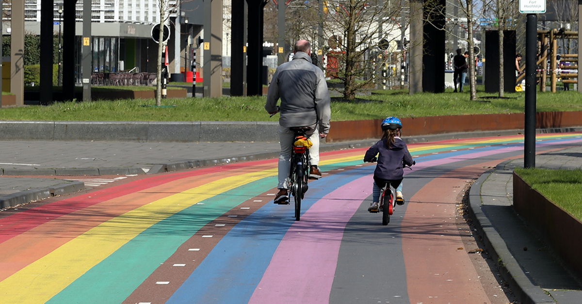 Isä ja lapsi pyöräilevät sateenkaaren värein maalatulla pyörätiellä.