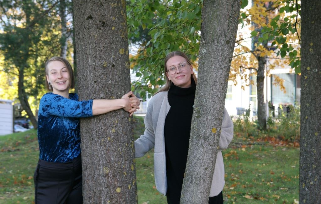 Kaksi naista seisoo ulkona ja toinen heistä halaa puuta.